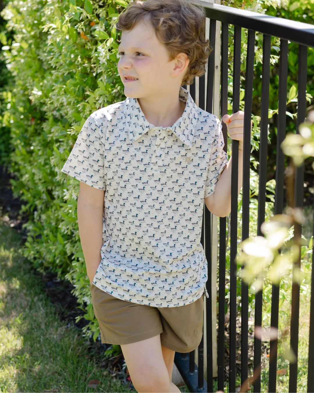 A young boy wearing a short-sleeve collared button-up shirt with a small repeating geometric pattern in beige and white, paired with brown shorts, standing outdoors next to a black metal fence, that store