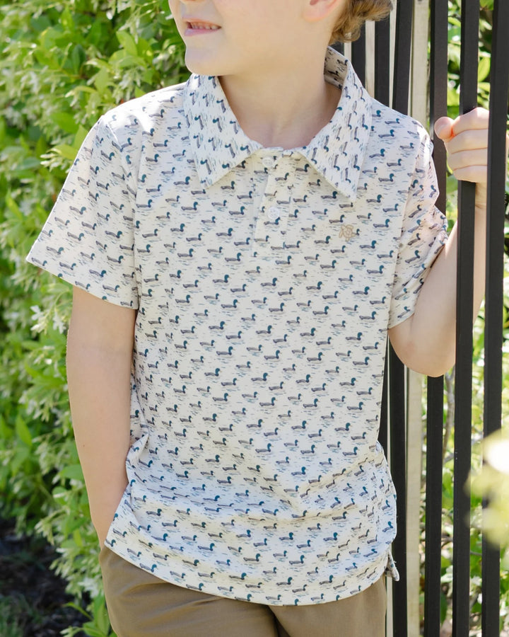 A child wearing a short-sleeve collared shirt with a small repeating blue and gray animal print on a light cream background, standing outdoors next to a black metal fence, that store