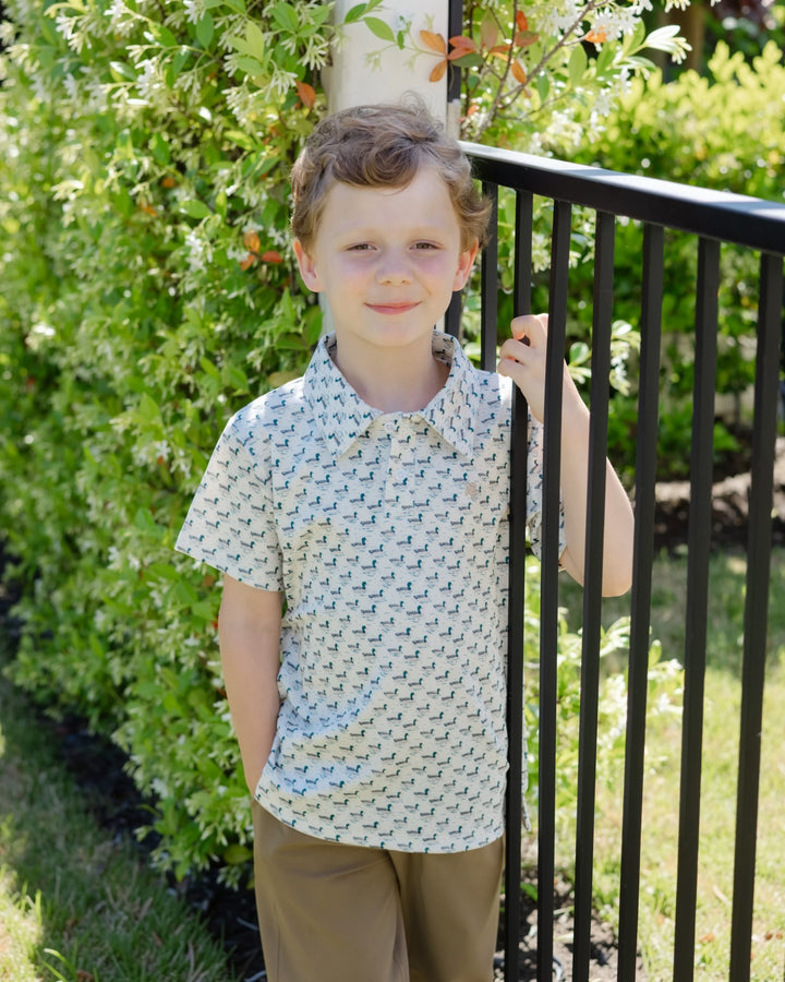 A young boy wearing a short-sleeve collared button-up shirt with a small repeating geometric pattern in light blue and white, paired with brown pants, standing outdoors next to a black metal fence with green bushes behind him, that store