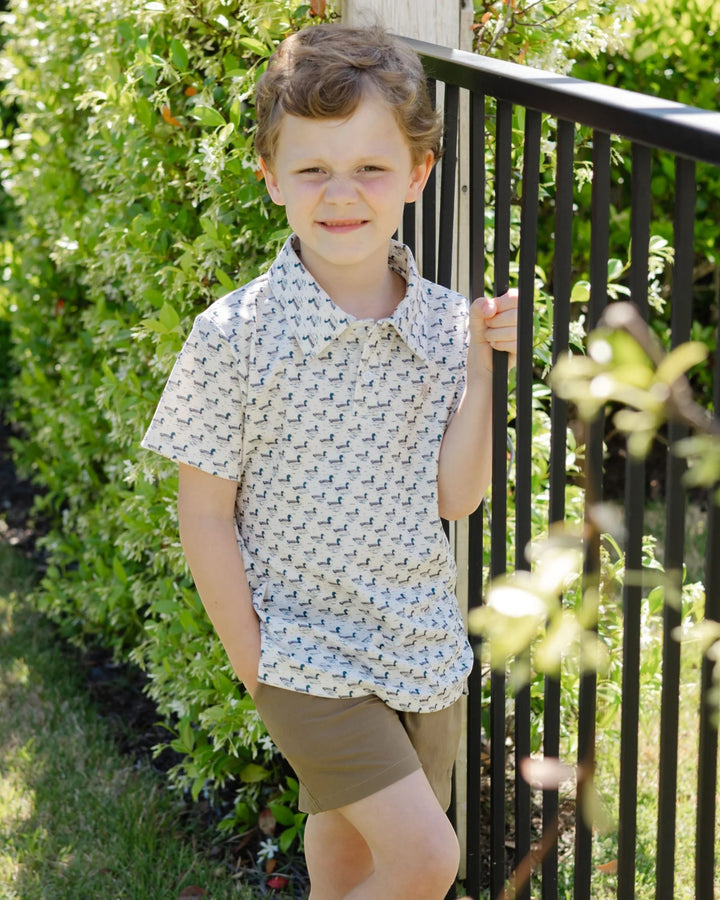A young boy wearing a short-sleeve button-up shirt with a small repeating pattern and brown shorts, standing outdoors next to a black metal fence with green bushes in the background, that store