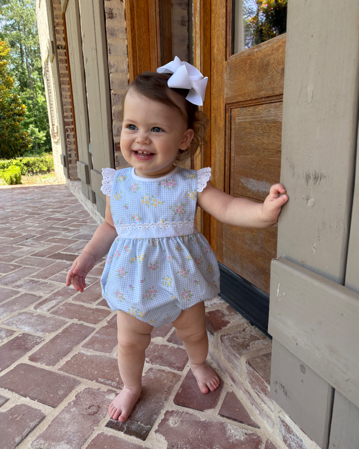 Baby in a light blue dress with floral patterns standing on a stone porch.