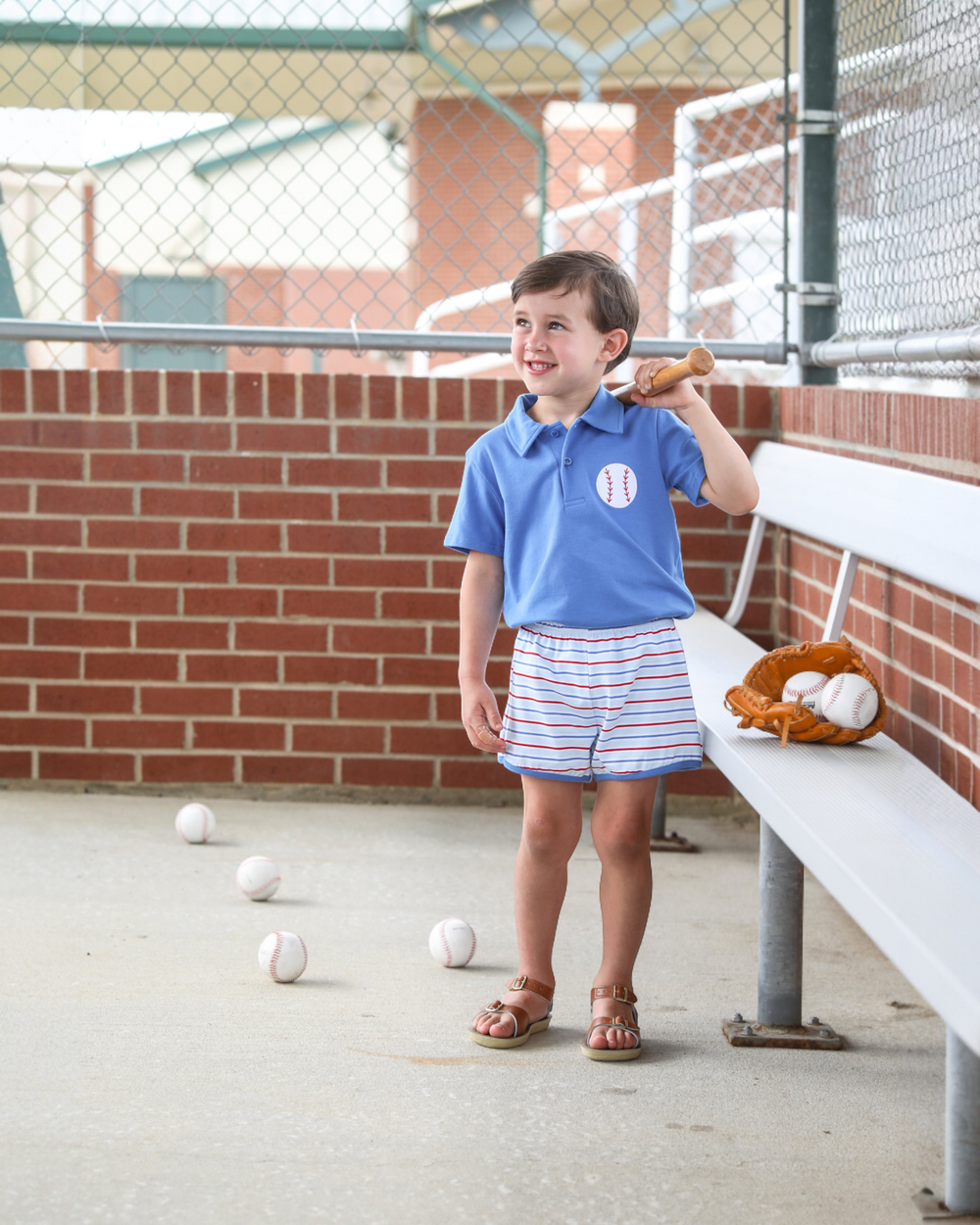 Young boy in a blue shirt and striped shorts standing in a batting cage with baseballs on the ground.