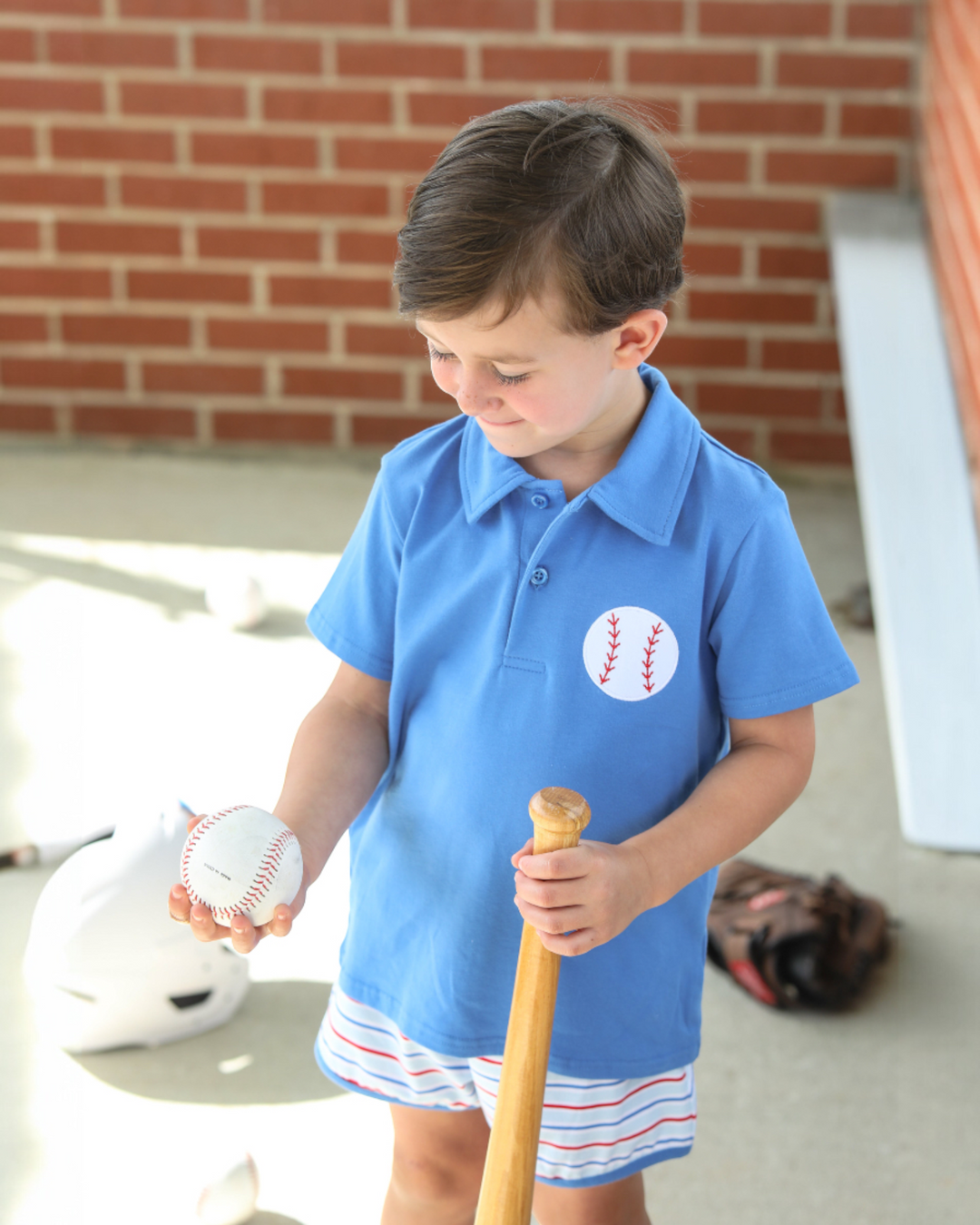 Child wearing a blue shirt with a baseball design, holding a bat and ball, standing in front of a brick wall.