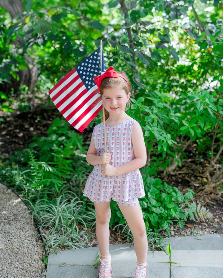 Young girl holding an American flag outdoors with greenery in the background, that store