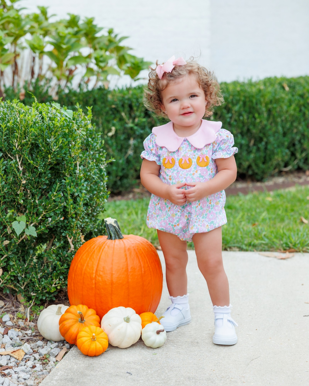 A toddler girl stands outdoors on a concrete path next to a large orange pumpkin and smaller pumpkins, wearing a short-sleeve floral romper with a pink Peter Pan collar and three orange jack-o&
