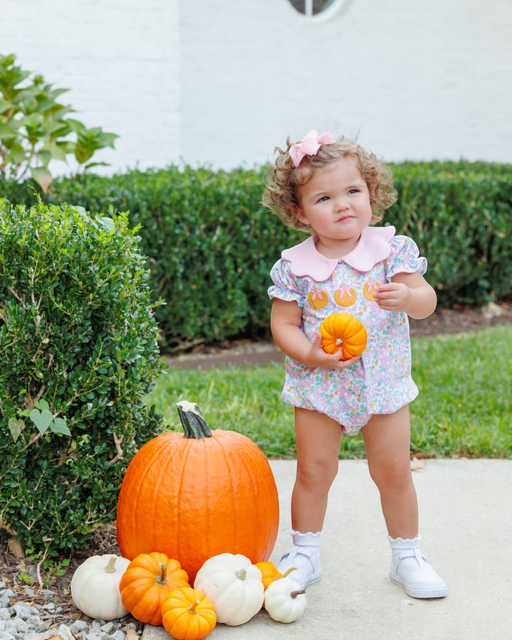 A toddler girl stands outdoors on a sidewalk next to a large orange pumpkin and smaller pumpkins, wearing a light floral romper with a pink collar and holding a small orange pumpkin, that store
