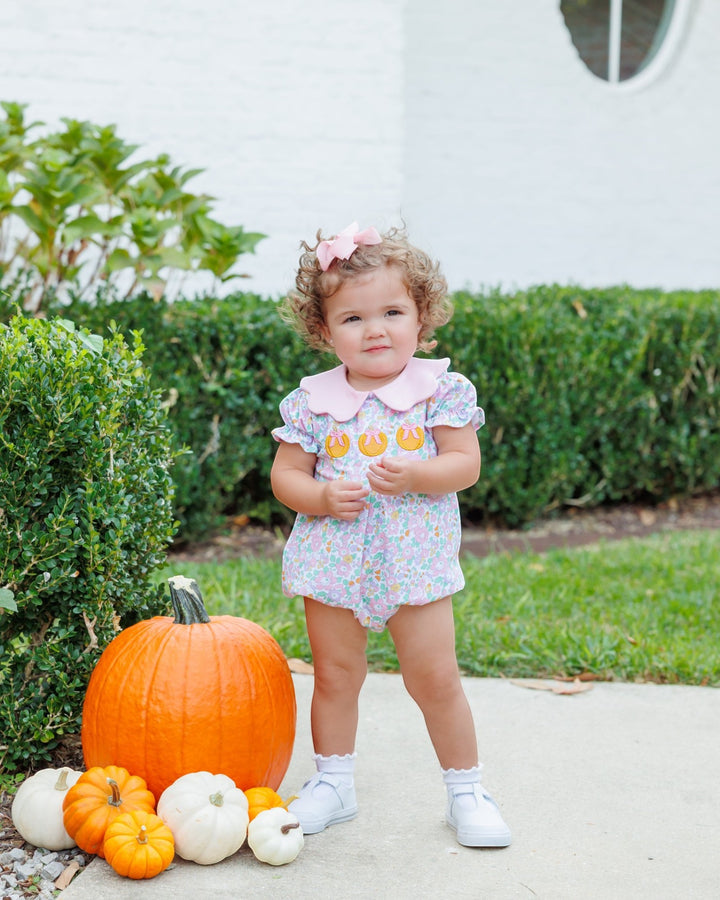A toddler girl stands outdoors on a concrete path wearing a light pink and multicolor floral romper with a pink collar and matching bow in her hair. She is surrounded by pumpkins and white houses in the background, that store