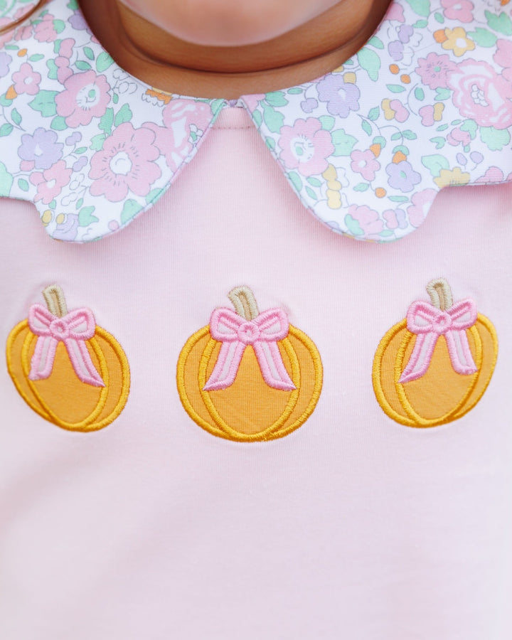 Close-up of a baby clothing detail showing a light pink top with three embroidered yellow pumpkins wearing pink bows, paired with a white floral Peter Pan collar, that store
