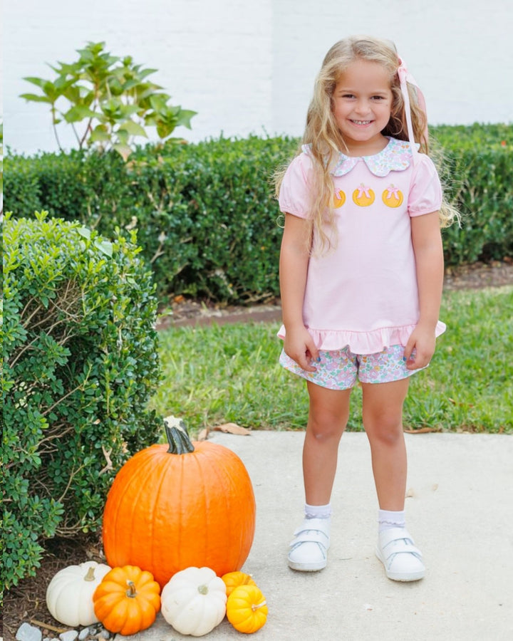 A young girl stands outdoors on a concrete path next to a large orange pumpkin and smaller pumpkins, wearing a light pink short-sleeve top with yellow sun graphics and patterned shorts, that store