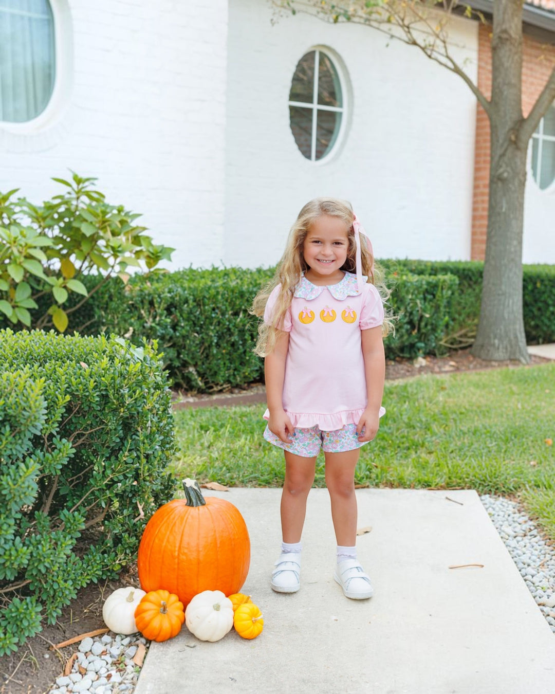 A young girl stands on a concrete path in front of a house, wearing a light pink short-sleeve top with three yellow pumpkin faces and gray patterned shorts. Large and small pumpkins are arranged on the path beside her, that store