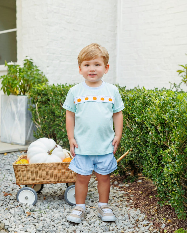 A young boy stands outdoors in front of a white brick wall and green hedges, wearing a light blue short-sleeve t-shirt with a row of orange pumpkin motifs across the chest, light blue shorts, and white sneakers. A wicker cart with white pumpkins sits to his left, that store