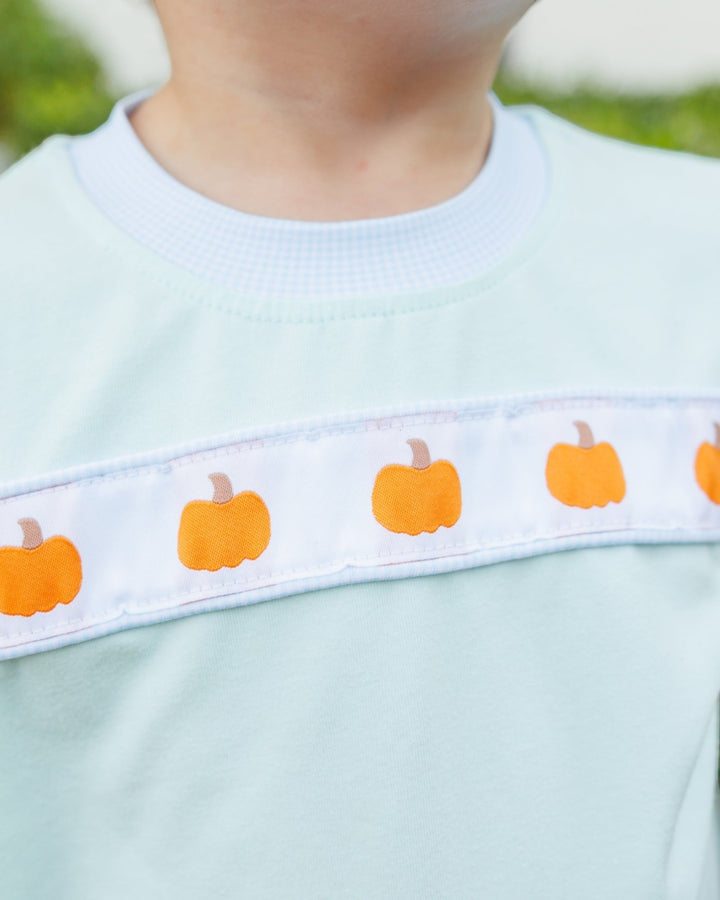 Close-up of a child wearing a pale mint green shirt with a horizontal white band across the chest featuring orange pumpkin illustrations, that store