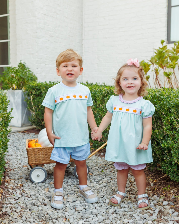 Two young children standing outdoors on a gravel path in front of a white brick wall and green hedges. The child on the left wears a light mint blue short-sleeve top and shorts, and the child on the right wears a matching light mint blue dress with a decorative orange and yellow band and pink shorts, that store