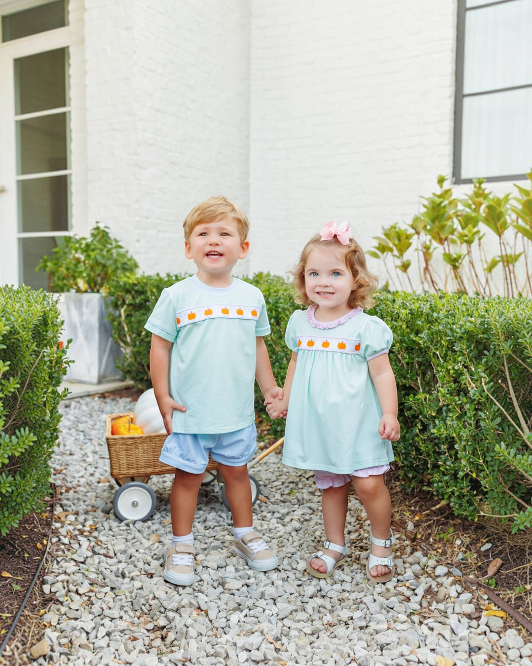 A young boy and girl stand on a gravel path outdoors in front of a white house, wearing matching light blue outfits with orange and yellow decorative trim across the chest, that store