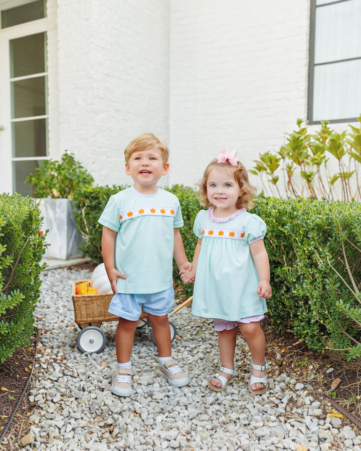 A young boy and girl stand on a gravel path outdoors in front of a white house, wearing matching light blue outfits with orange and yellow decorative trim across the chest, that store