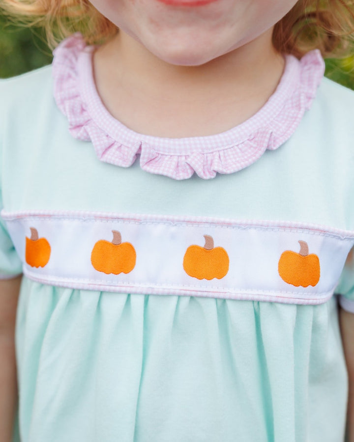 Close-up of a child wearing a light mint green dress with a pink gingham ruffled collar and a white smocked band featuring orange pumpkin motifs, that store