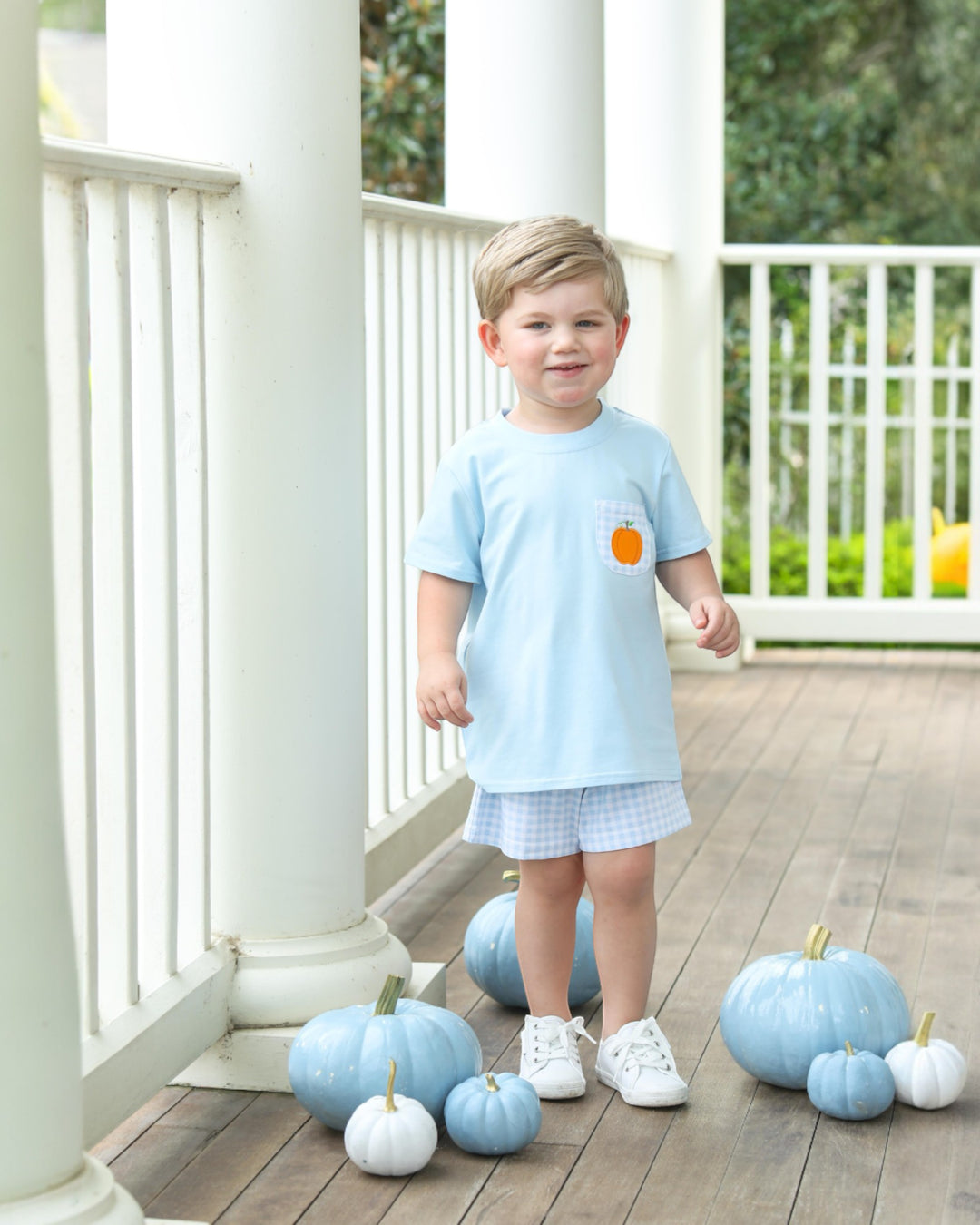 Child in light blue shirt with orange patch standing on a wooden deck with decorated pumpkins.