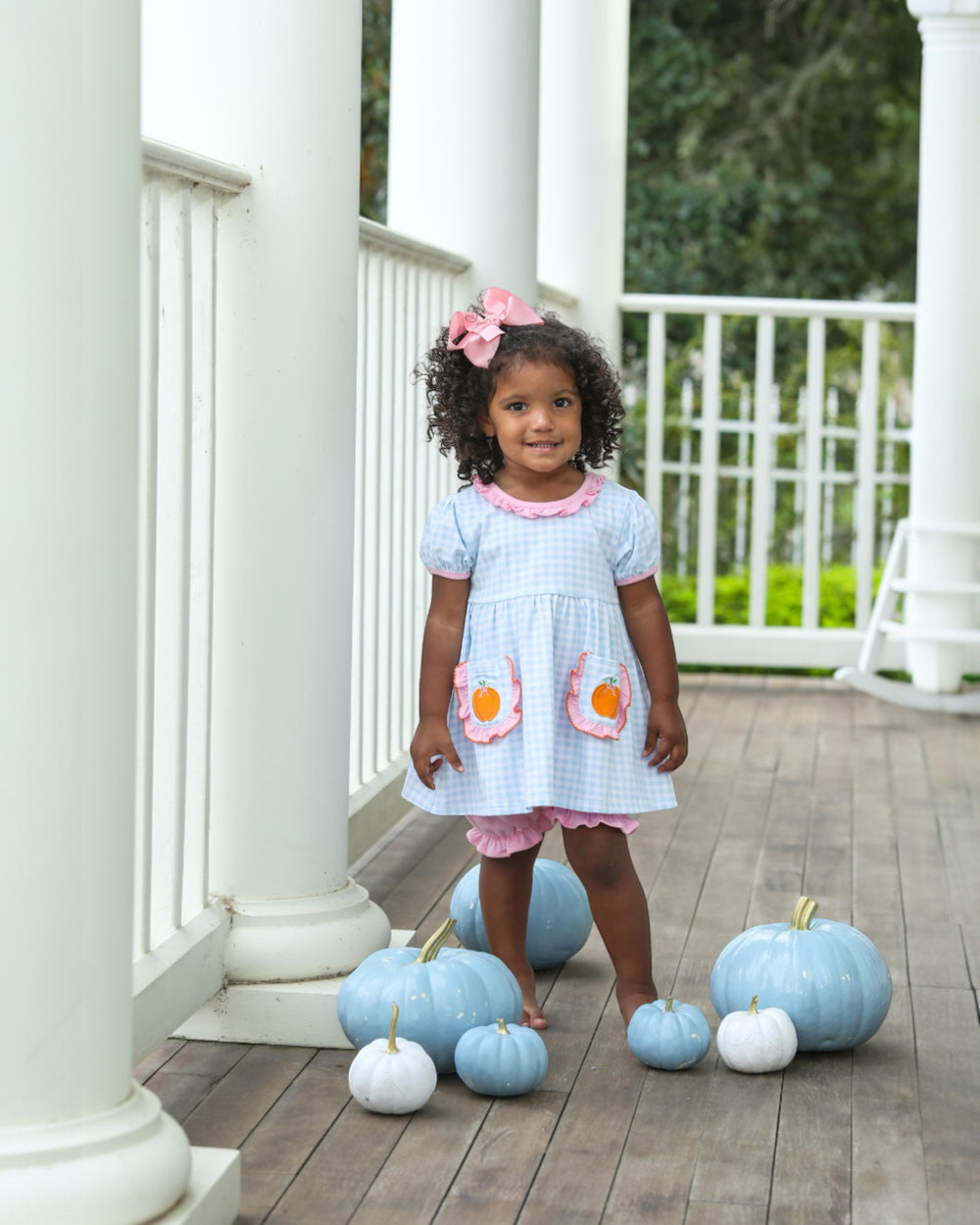 Young girl in a light blue dress with pink accents standing on a wooden porch with pastel pumpkins.