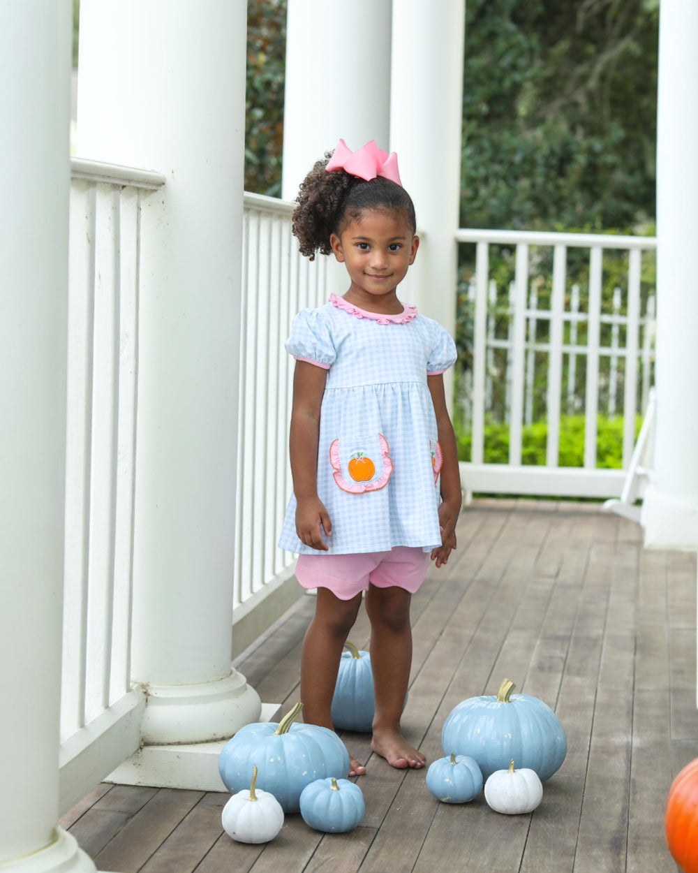 Young girl in a light blue dress with pink trim standing on a wooden deck with blue and white pumpkins around her.