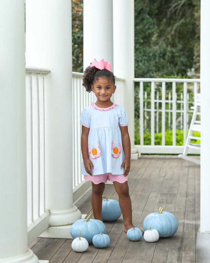 Young girl in a light blue dress with pink accents standing on a wooden deck with pumpkins.