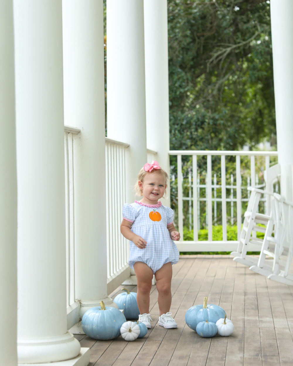 Child in a light blue dress standing on a wooden porch with decorative pumpkins.