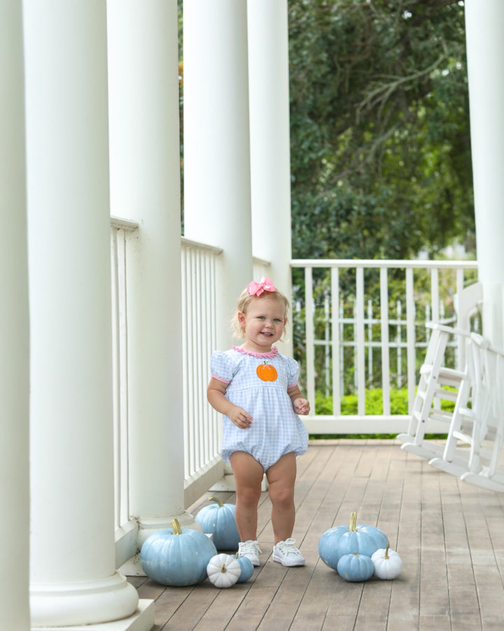 Child in a light blue dress standing on a wooden porch with decorative pumpkins.