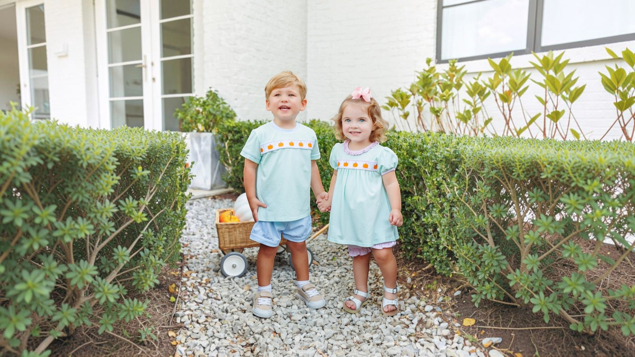 Two toddlers standing on a gravel path between green hedges in front of a white house. The boy wears a light blue t-shirt with yellow-orange embroidered dots and denim shorts, while the girl wears a matching light blue dress, that store