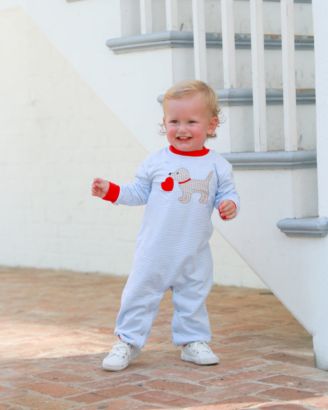 Baby wearing a white onesie with red and gray designs, standing on a wooden floor.