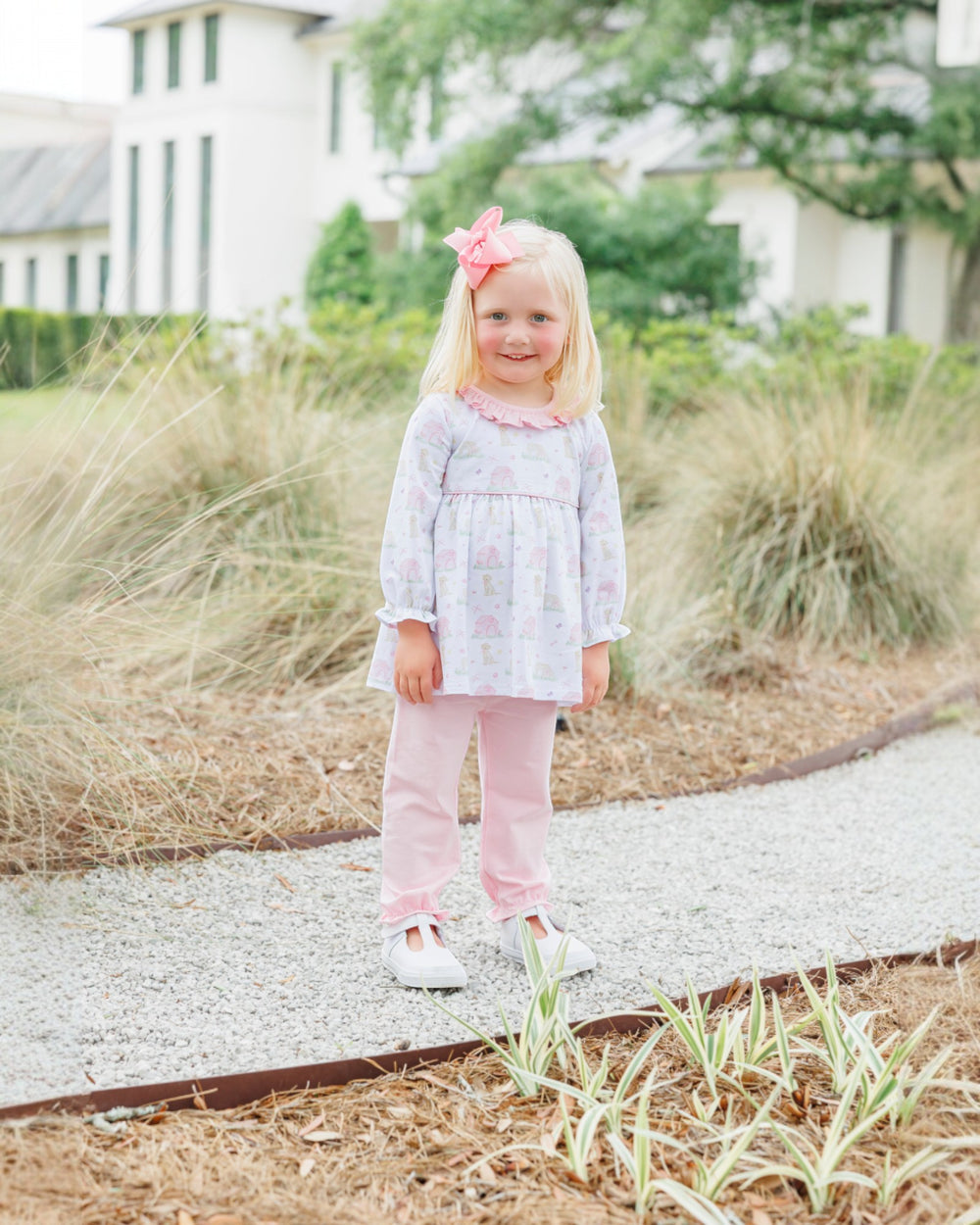 Young girl in a pink outfit standing outdoors with grass and a house in the background