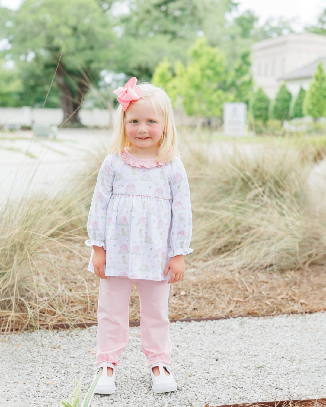 Young girl in a light purple floral dress and pink pants standing outdoors with greenery in the background.