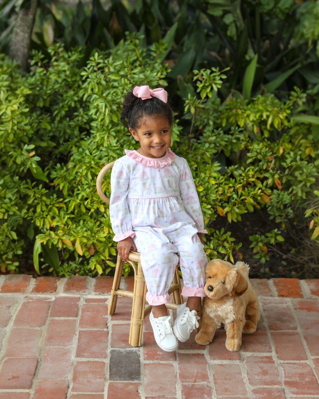 Young girl in a light outfit sitting on a stool with a teddy bear outdoors.