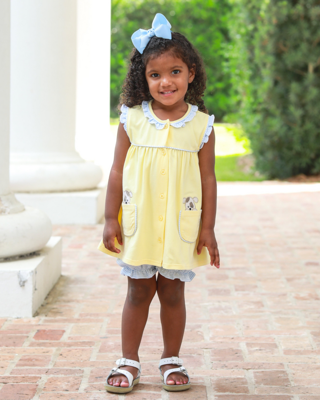 Young girl in a yellow dress with a blue bow standing outdoors on a brick path.