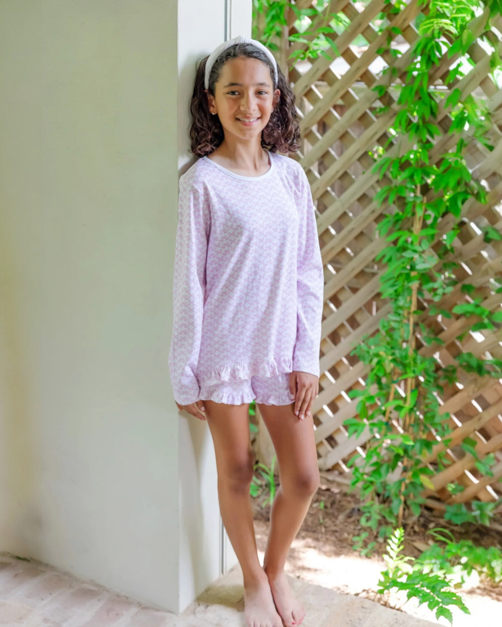 Young girl wearing a light purple pajama set standing against a white wall with greenery in the background.
