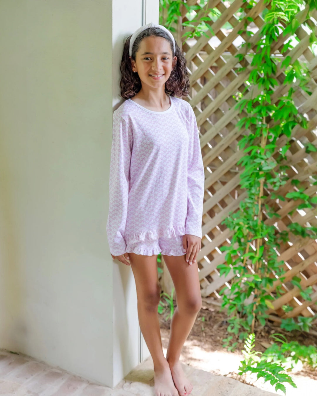 Young girl wearing a light purple pajama set standing against a white wall with greenery in the background.