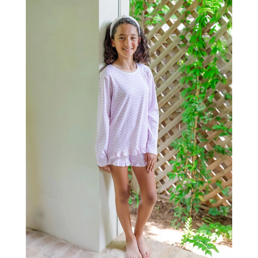 Young girl wearing a light purple pajama set standing against a white wall with greenery in the background.