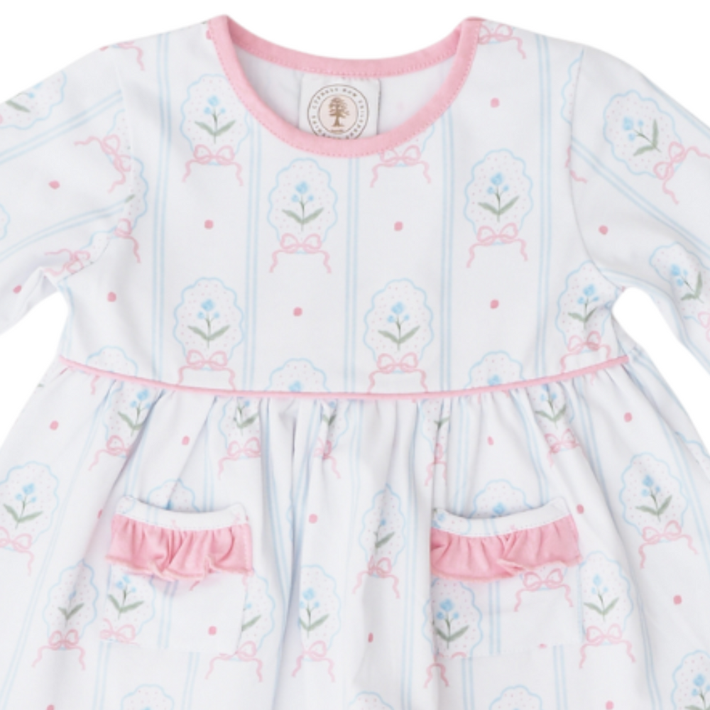 A baby girl's long-sleeve dress with a white base, light blue vertical stripes, and a pink and blue floral bow pattern. The dress features a pink crew neck, a pink waistband, and two ruffled pocket flaps at the skirt, that store