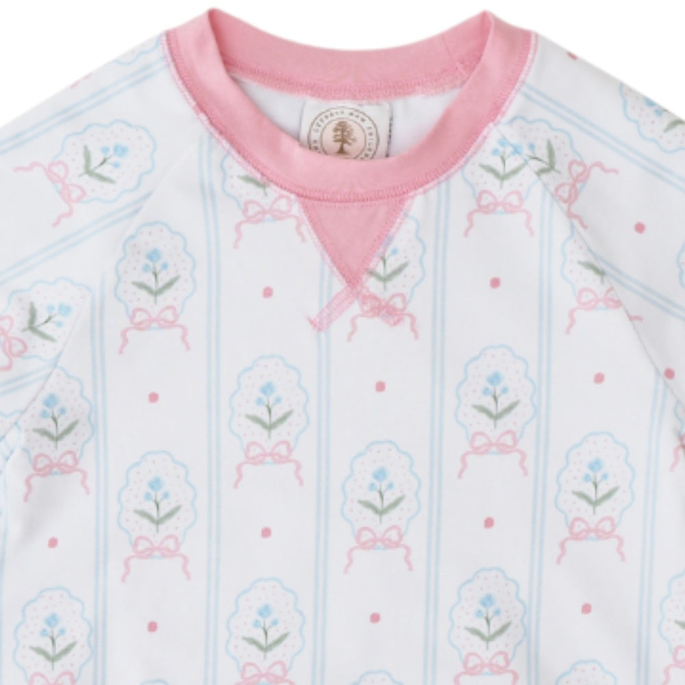 A close-up of a baby long-sleeve top featuring a white base with light blue vertical stripes, pink bow details, and small floral motifs in pink, blue, and green. It has a pink crew neck and a small round brand label inside the collar, that store