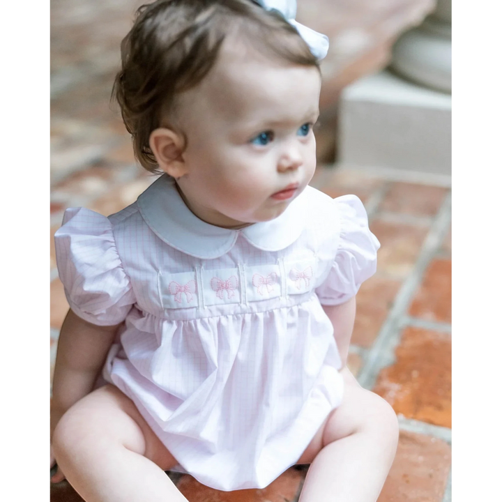 Baby wearing a white dress with ruffled sleeves sitting on a tiled floor.