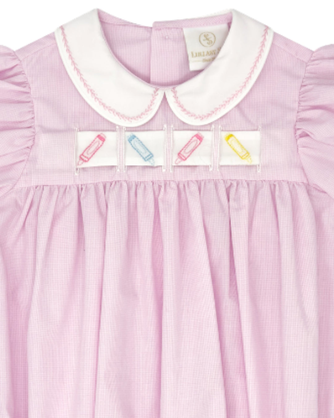 A close-up of a light pink gingham nightgown featuring a white Peter Pan collar with pink piping, smocked detailing, and four embroidered patch pockets with colorful crayon designs, that store