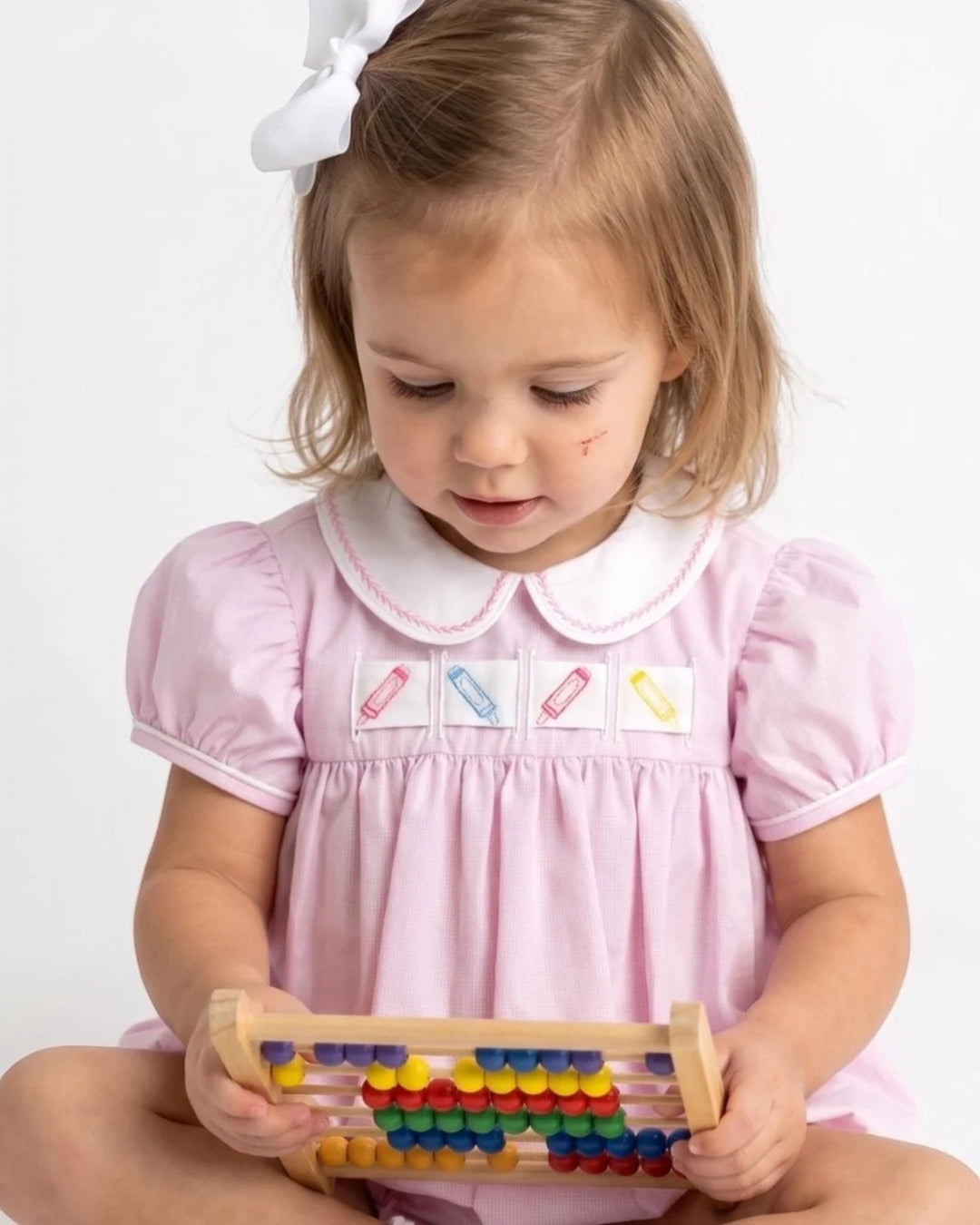 A young toddler girl in a light pink dress with a white Peter Pan collar holds a wooden abacus toy against a plain white background, that store