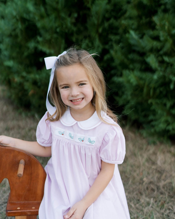 A young girl with blonde hair wearing a light pink smocked dress with a white Peter Pan collar, a white bow headband, and a white bow, sitting on a wooden horse outdoors that store