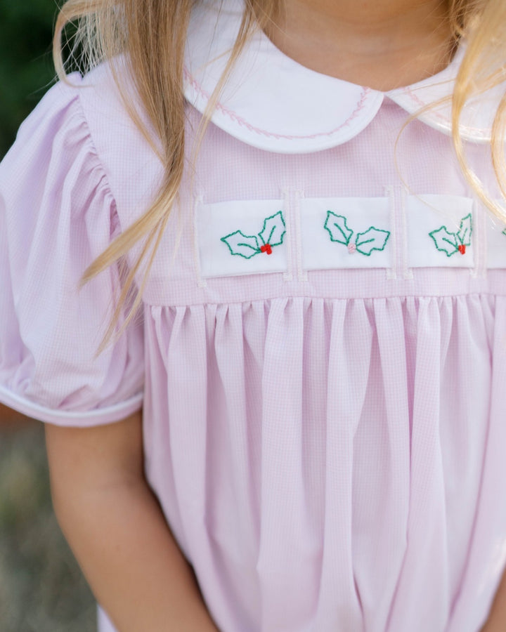 Close-up of a child wearing a light pink smocked dress with a white Peter Pan collar and embroidered holly motifs on the chest, that store