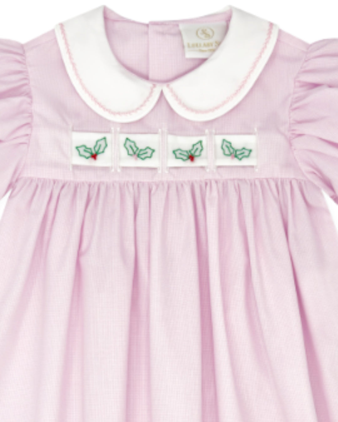 A close-up of a light pink baby dress with a white Peter Pan collar, featuring four embroidered holly motifs across the chest, that store
