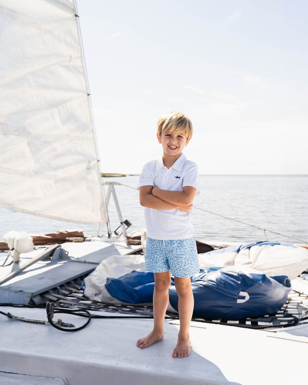 Young boy standing on a sailboat with arms crossed, wearing a white shirt and patterned shorts.
