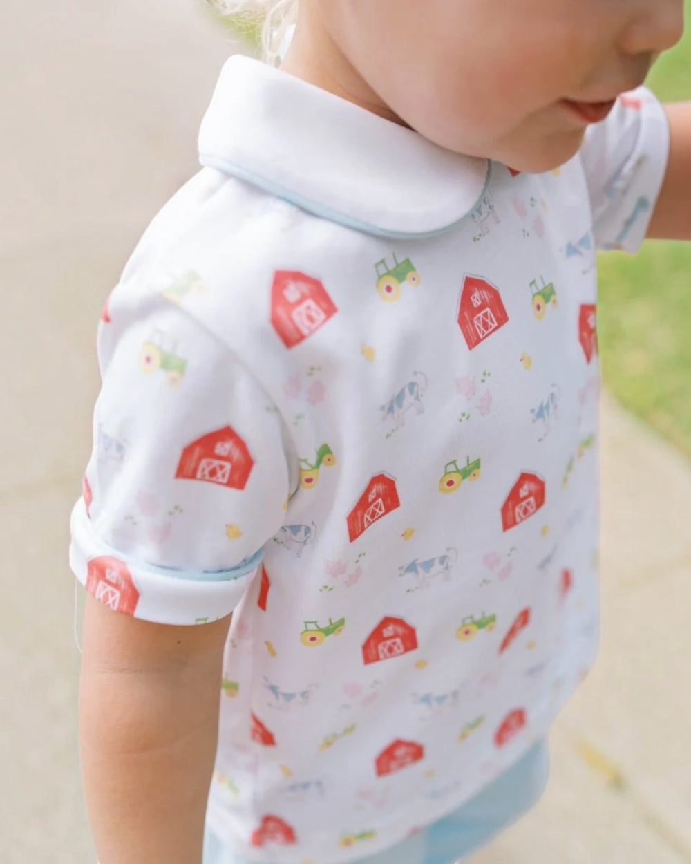 A close-up of a toddler wearing a white short-sleeve polo shirt with a colorful farm-themed print of red barns, green tractors, and yellow tractors, featuring a white collar, that store