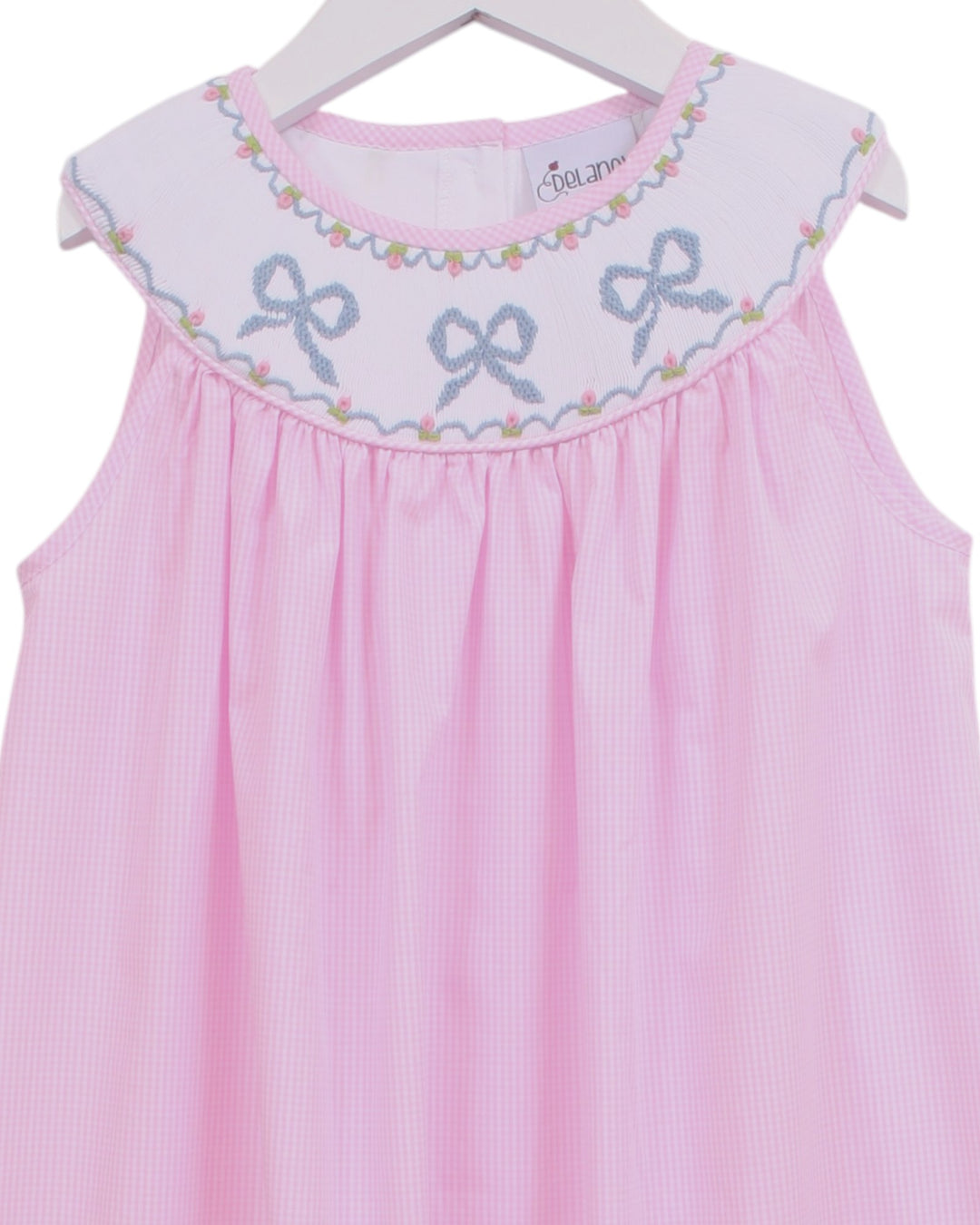 Pink gingham sleeveless baby dress with a white embroidered yoke featuring blue bows and a floral border, hanging on a white hanger, that store
