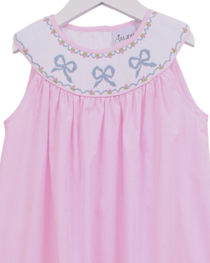 Pink gingham sleeveless baby dress with a white embroidered yoke featuring blue bows and a floral border, hanging on a white hanger, that store