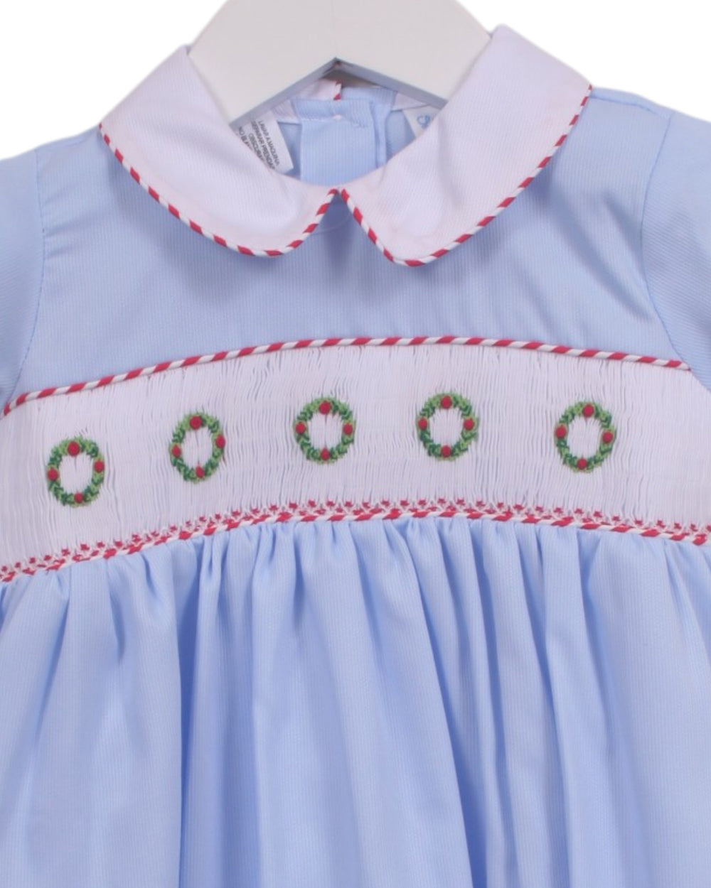 A light blue baby dress with a white Peter Pan collar and smocked chest detailing featuring red, white, and green wreaths, shown on a white hanger, that store