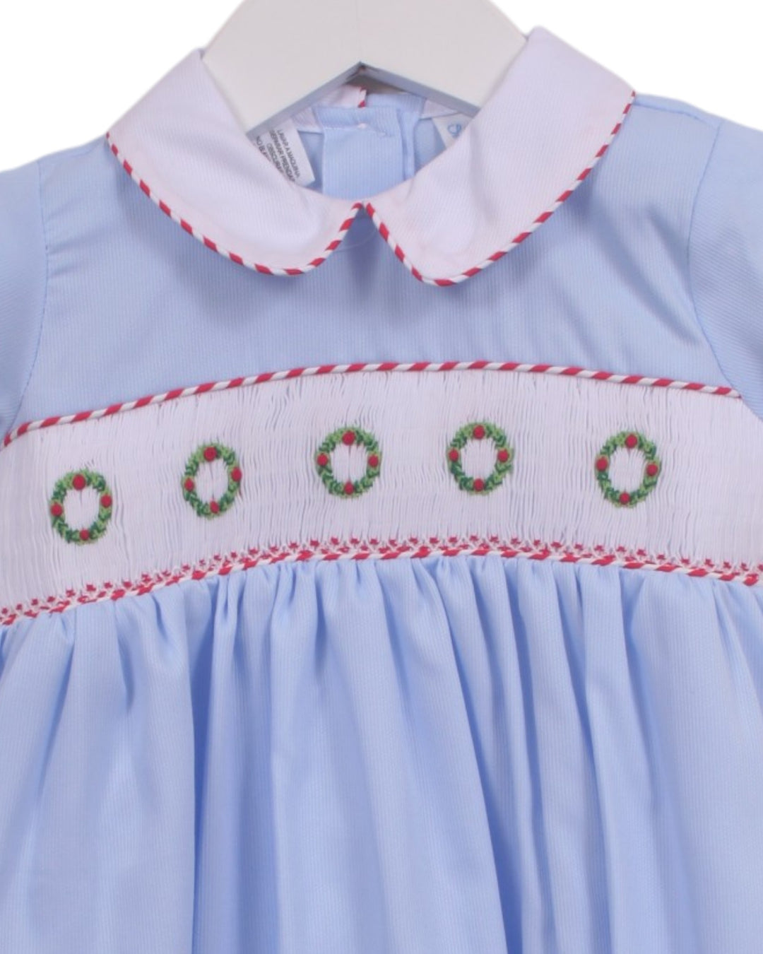 A light blue baby dress with a white Peter Pan collar and smocked chest detailing featuring red, white, and green wreaths, shown on a white hanger, that store
