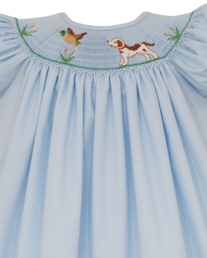 A light blue baby smocked dress featuring embroidered details of a brown and white dog and a duck at the neckline, set against a plain white background, that store